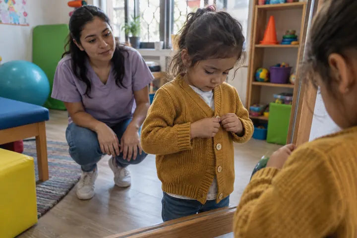 Una niña se abrocha sola los botones de su suéter frente al espejo